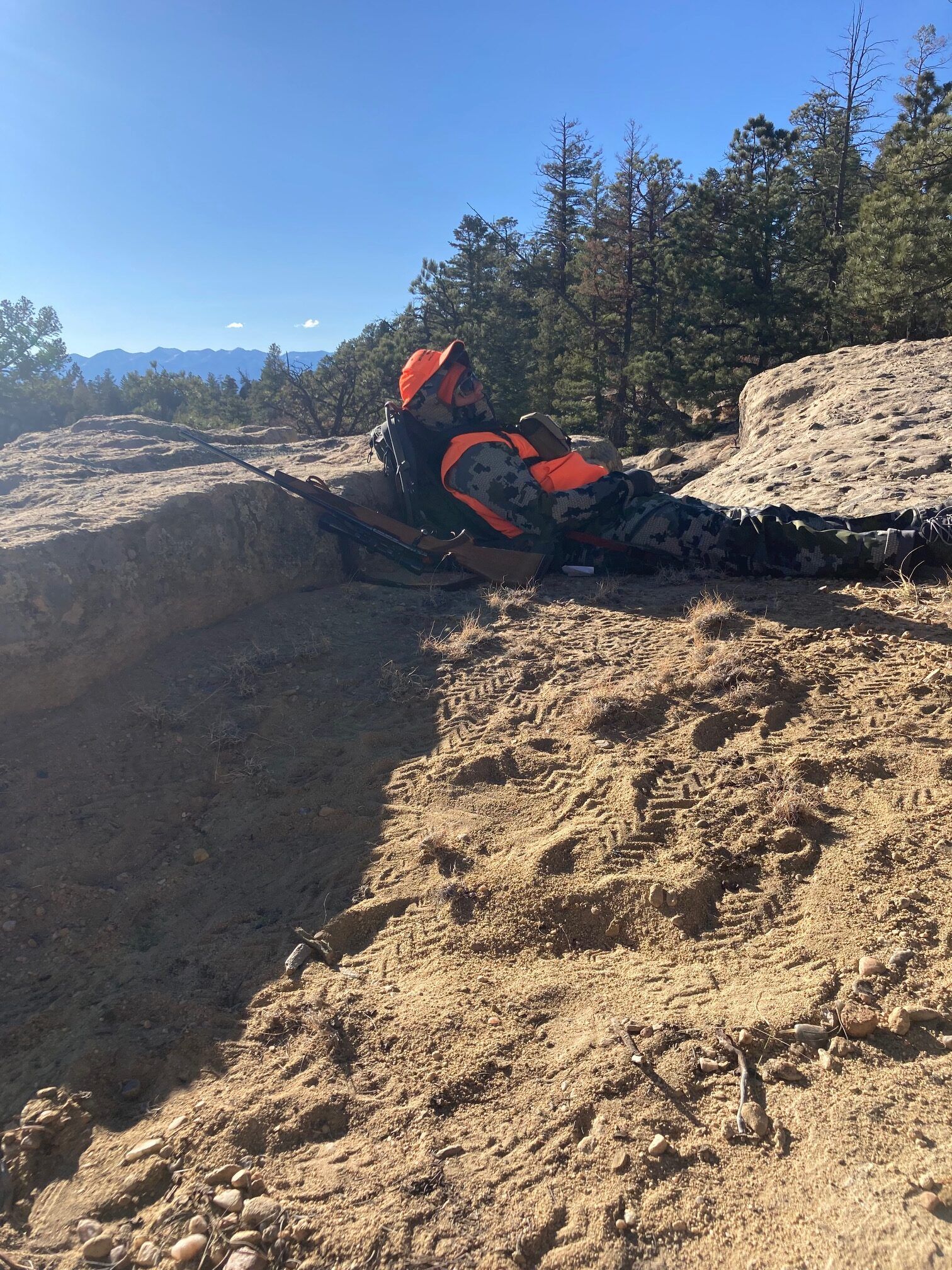 A hunter in camouflage and orange gear sitting in a mountainous area.