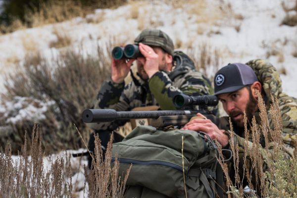 Two hunters using binoculars and rifle in snowy terrain.