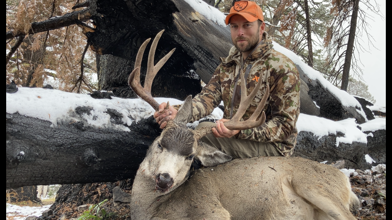 Hunter posing with a mule deer buck in a snowy forest setting.
