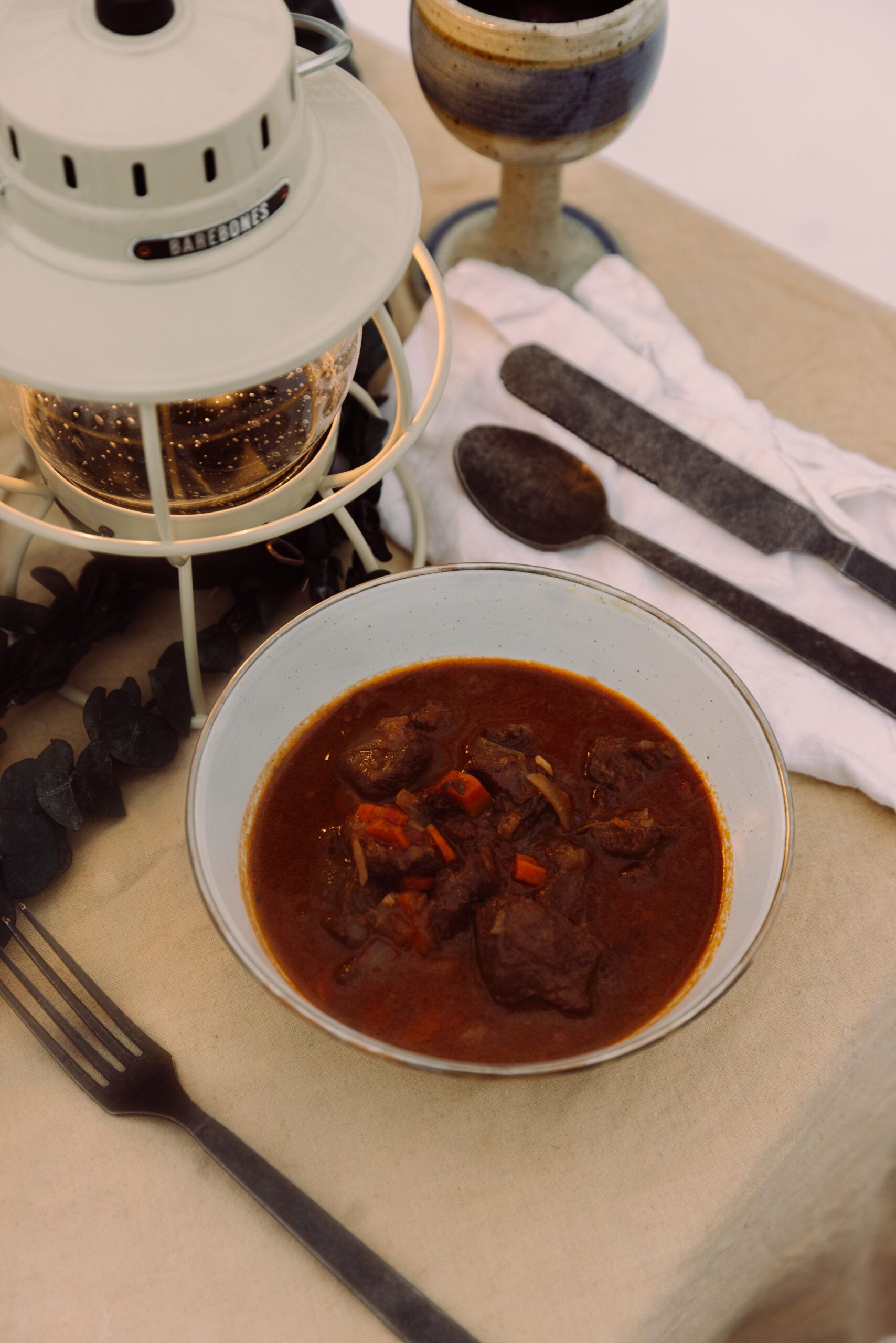 Bowl of deer meat stew with lantern and utensils in camp setting.