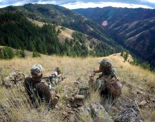 two hunters resting on a grassy hill overlooking mountain landscape