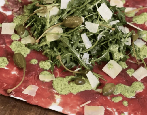Plate of venison carpaccio with arugula and capers on a wooden table.