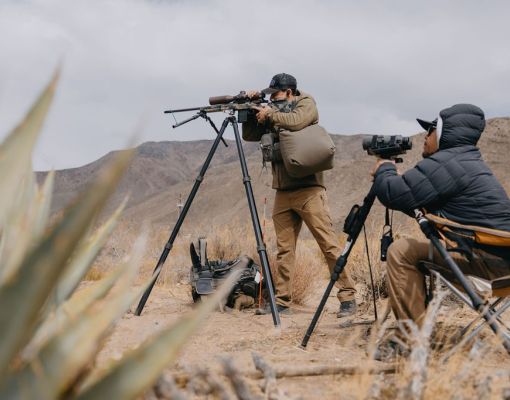Two individuals observing wildlife with rifles and spotting scopes in desert setting.
