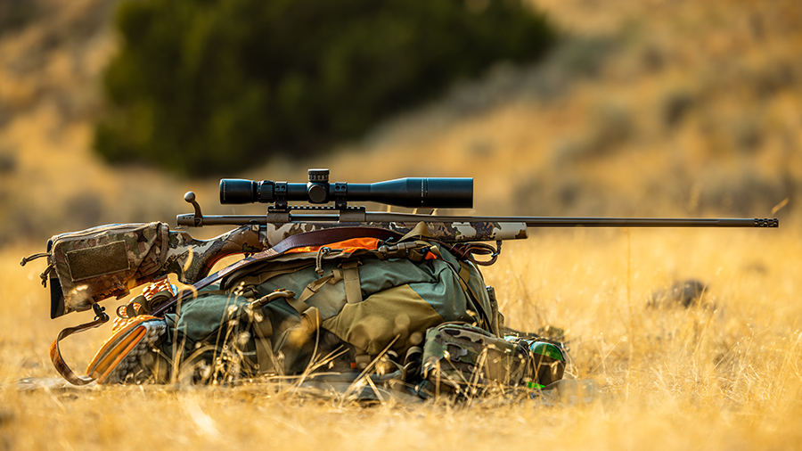 Hunting rifle resting on a backpack in a dry grassy area.