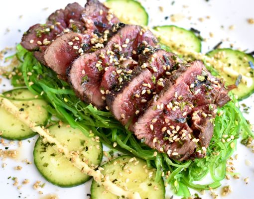 Grilled mule deer steak on a plate with cucumbers and seaweed salad.