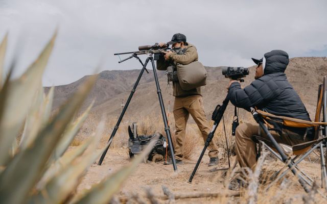Two individuals observing wildlife with rifles and spotting scopes in desert setting.