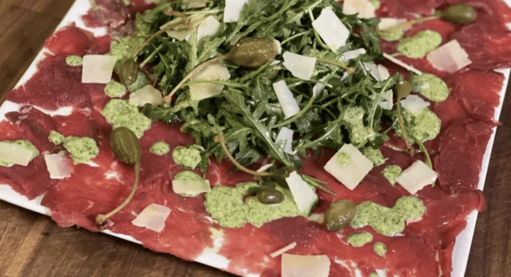 Plate of venison carpaccio with arugula and capers on a wooden table.