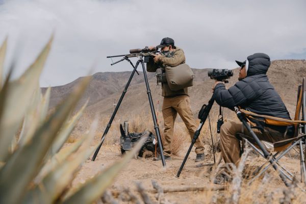 Two individuals observing wildlife with rifles and spotting scopes in desert setting.