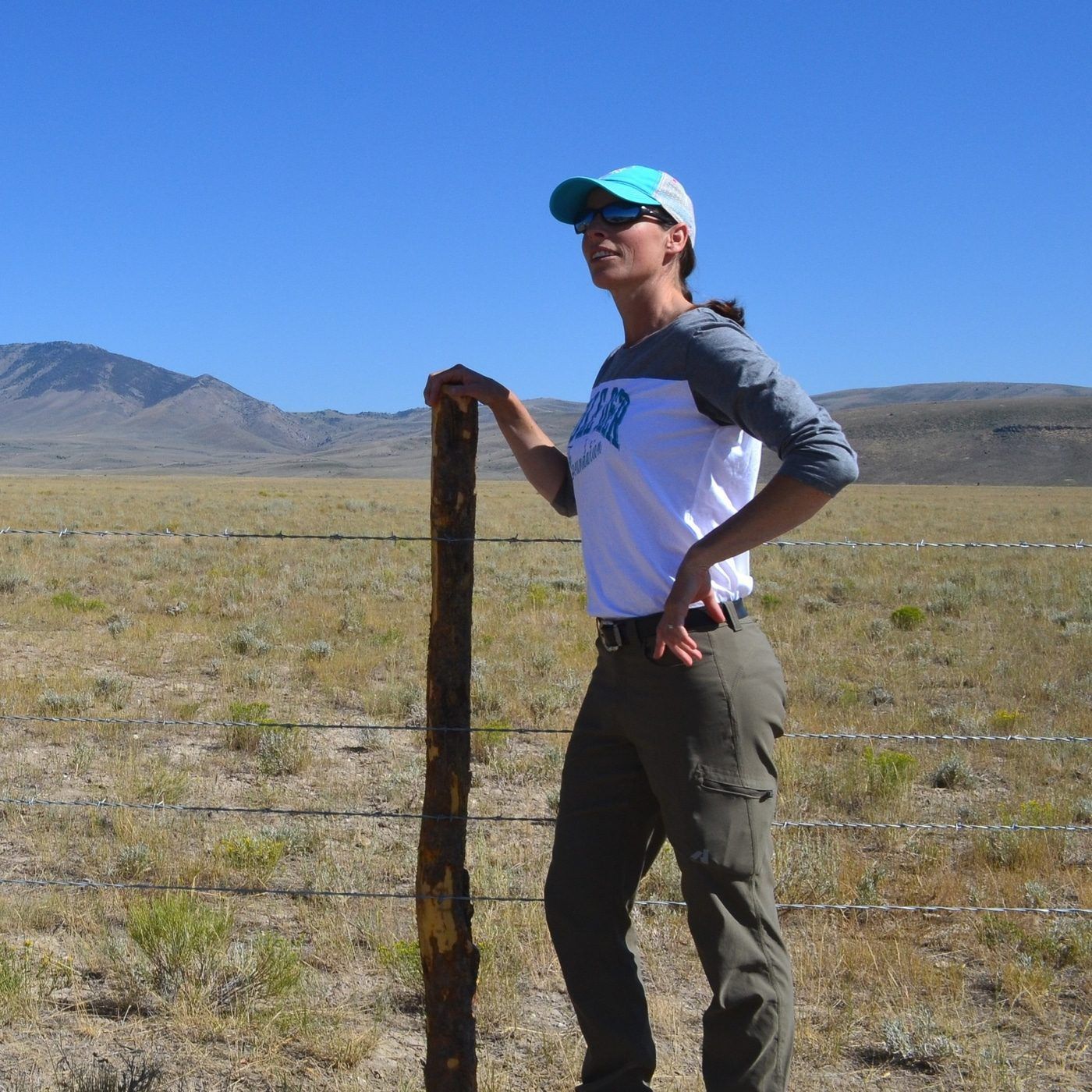 Woman standing by a fence in a grassy landscape under a clear blue sky.