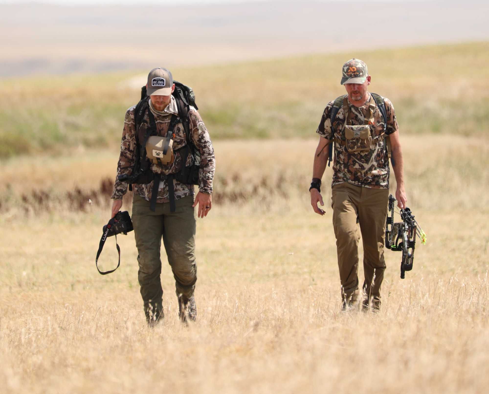 Two hunters walking in a grassy field, one with a camera, one with a bow
