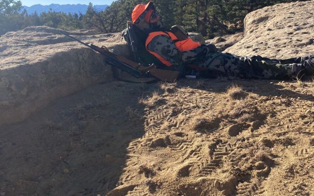 A hunter in camouflage and orange gear sitting in a mountainous area.