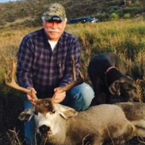 Man with mule deer in field with dog nearby.