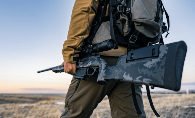 Hunter with rifle in a field during sunset.