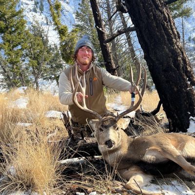 Hunter poses with harvested mule deer buck in snowy forest setting.