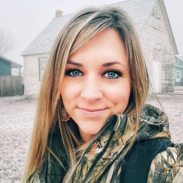Woman in camouflage jacket smiling outside a house.