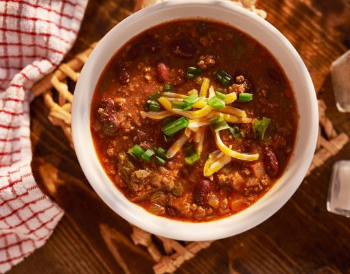 Bowl of chili topped with cheese and green onions on wooden table.