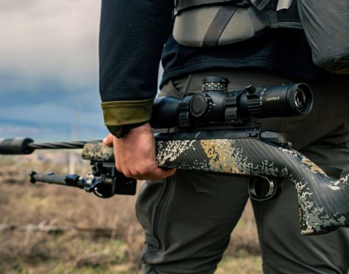 A hunter holding a rifle in a grassy landscape under a cloudy sky.