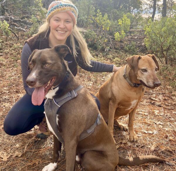Woman posing with two dogs in a forest setting.