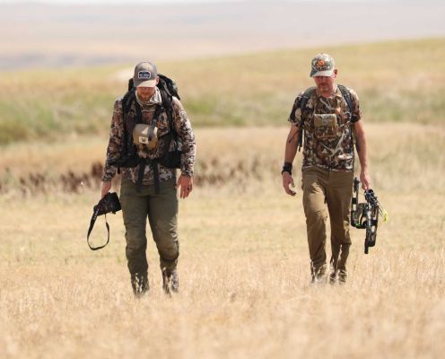 Two hunters walking in a grassy field, one with a camera, one with a bow
