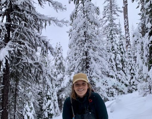 Person smiling in a snowy forest setting with evergreen trees.