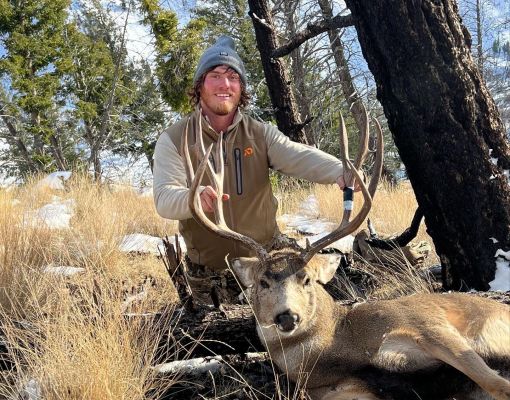 Hunter poses with harvested mule deer buck in snowy forest setting.
