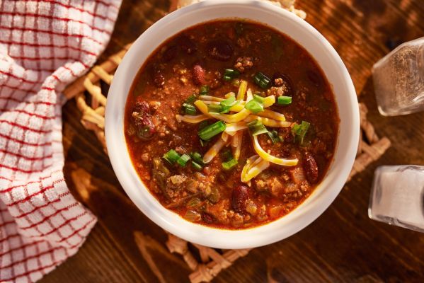Bowl of chili topped with cheese and green onions on wooden table.