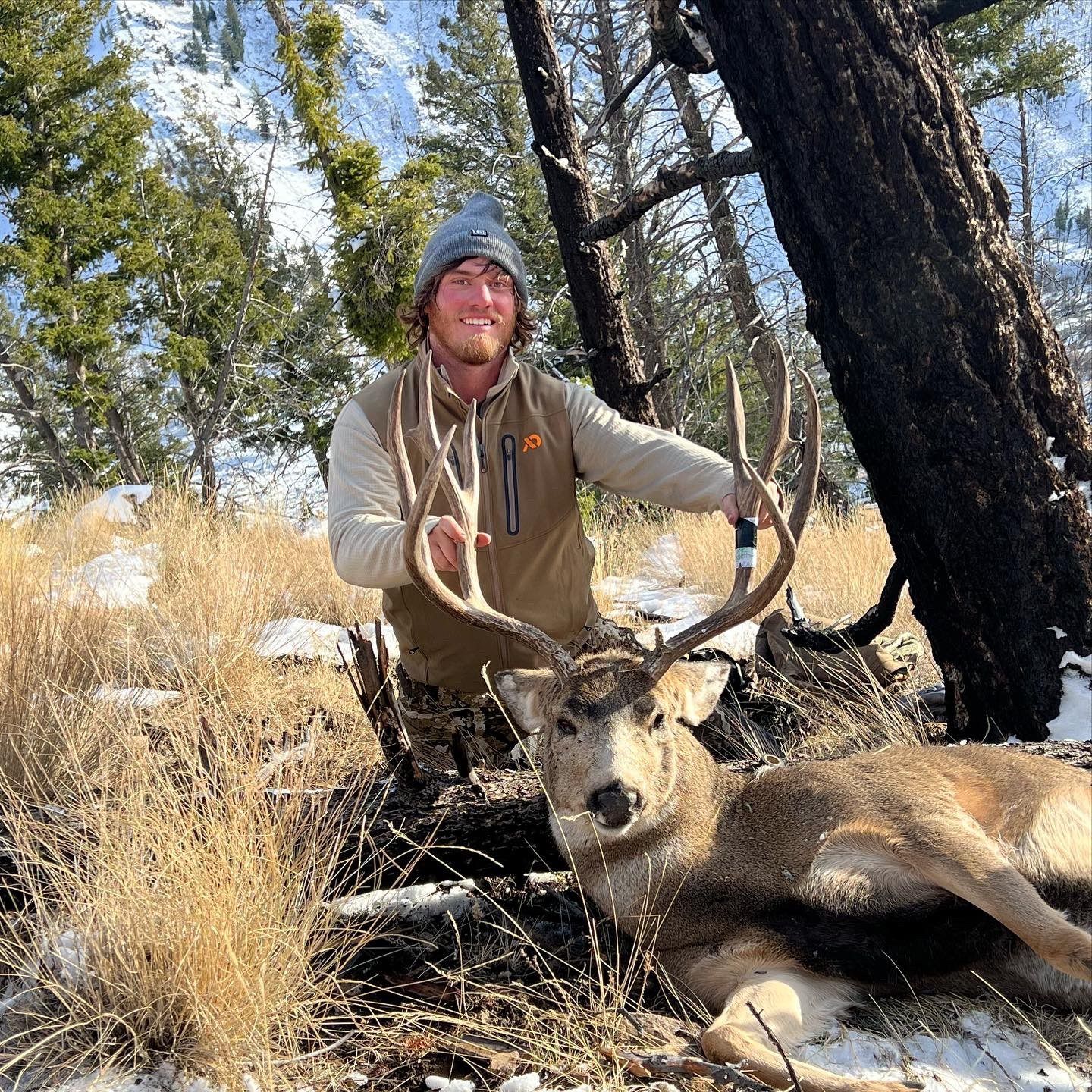 Hunter poses with harvested mule deer buck in snowy forest setting.