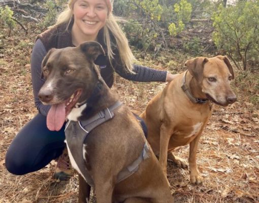 Woman posing with two dogs in a forest setting.