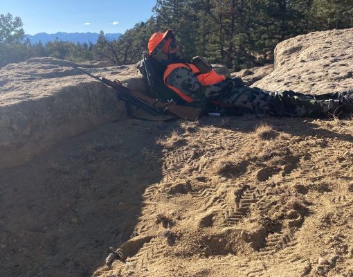 A hunter in camouflage and orange gear sitting in a mountainous area.