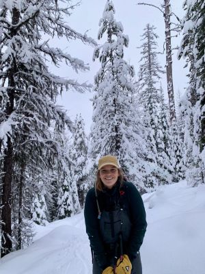 Person smiling in a snowy forest setting with evergreen trees.