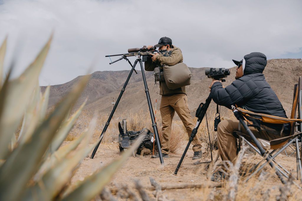 Two individuals observing wildlife with rifles and spotting scopes in desert setting.