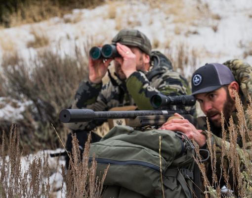 Two hunters using binoculars and rifle in snowy terrain.
