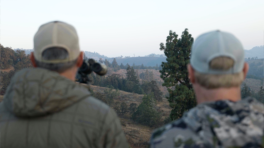 Two hunters observing a distant mountain landscape with binoculars.