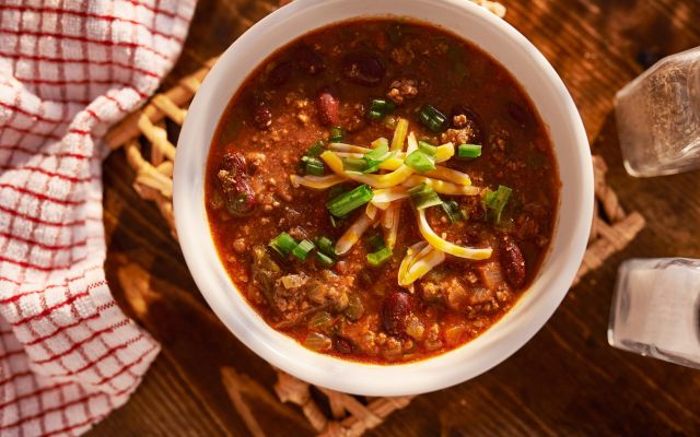 Bowl of chili topped with cheese and green onions on wooden table.