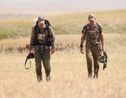 Two hunters walking in a grassy field, one with a camera, one with a bow