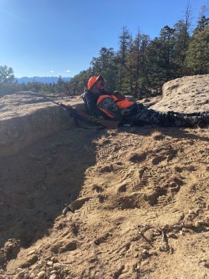 A hunter in camouflage and orange gear sitting in a mountainous area.