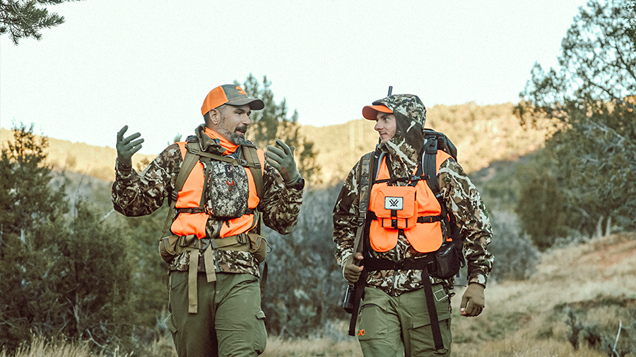 Two hunters wearing camouflage gear and orange safety vests talking on a trail