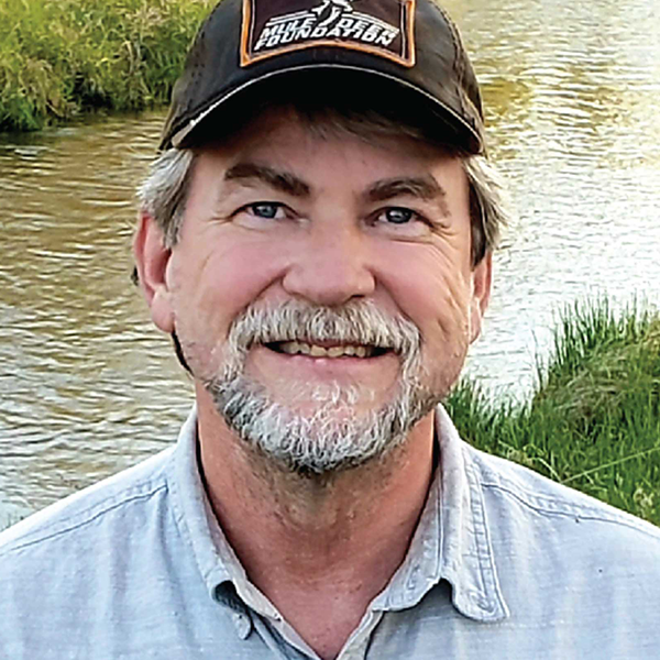 A man smiling by a river with grass in the background.