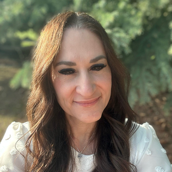 Woman smiling outdoors with greenery in the background.