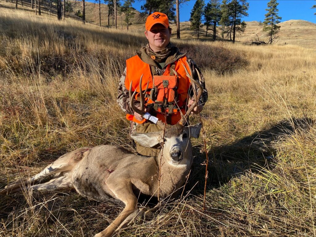 Hunter in orange vest poses with a mule deer buck on a grassy hillside.
