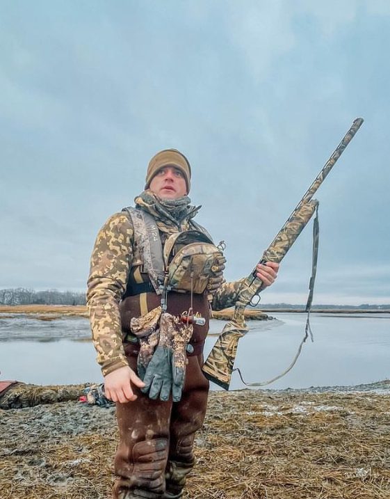 Hunting scene by riverbank with a man holding a rifle in camouflage.