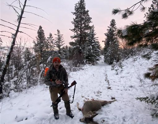 Hunter standing by mule deer in snowy forested area.
