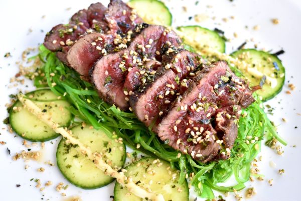 Grilled mule deer steak on a plate with cucumbers and seaweed salad.