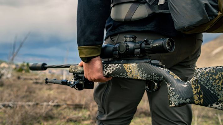 A hunter holding a rifle in a grassy landscape under a cloudy sky.